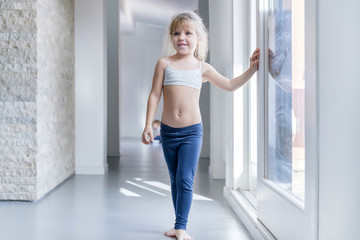 Fototapeta na wymiar Little blond girl in white top and blue leggings standing in a room near the window