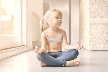 Little blond girl in white top and blue leggings doing a yoga exercise in the sunny room