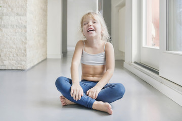 Smiling little blond girl in white top and blue leggings sitting on the floor in a room with crossed legs