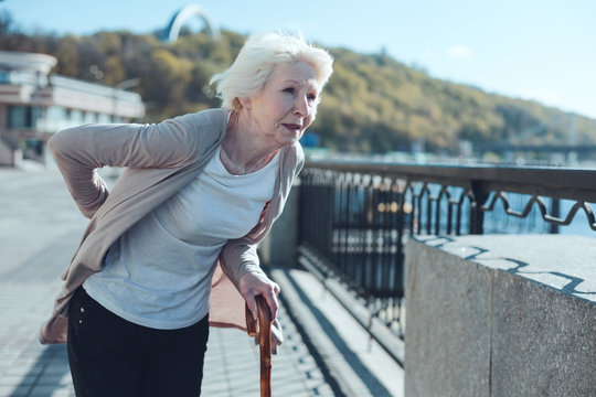 Physically Exhausted Elderly Woman Leaning On A Cane And Looking Into Vacancy While Struggling From A Physical Discomfort In A Low Back Zone.