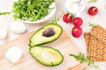 Green ripe avocado on wooden chopping board. Red garden radish, arugula, eggs and rye bread crusts. Healthy food