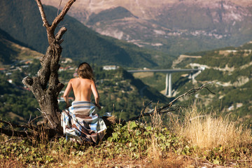 A naked girl in a plaid sitting on a tree with a cup of coffee and looking at the rural city. A...