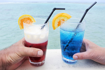Man and woman holds a red and blue cocktail drinks with ice served over a lagoon of a tropical island resort