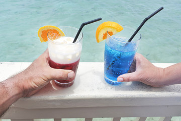 Man and woman holds a red and blue cocktail drinks with ice served over a lagoon of a tropical island resort