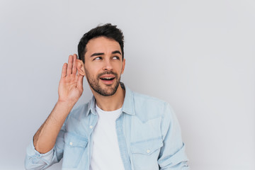 Portrait of handsome interested male placing hand on ear asking someone to speak up, isolated over white background. Young stylish man which overhears conversation in the studio. Copy space for text.