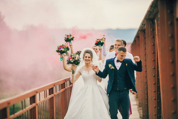 Newlyweds and their friends running with colors smoke bombs at old stone bridge. Happy wedding...