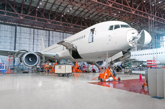 Passenger airplane on maintenance of engine and fuselage repair in airport hangar. Aircraft with open hood on the nose and engines, as well as the luggage compartment.