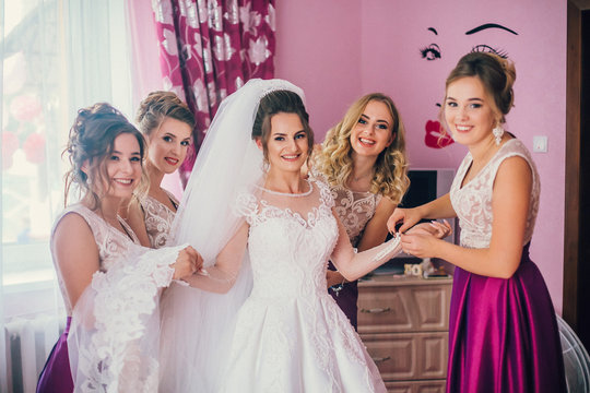 Beautiful bridesmaids help put on wedding dress on gorgeous blonde bride in luxury hotel room morning before the wedding