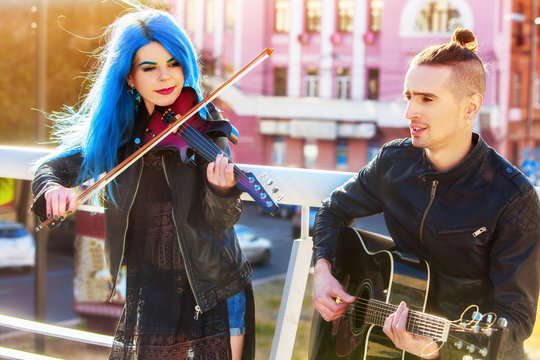 Playing viola woman and man perform music on violin and guitar in city outdoor. Girl with blue hairstyle and eyebrows performing jazz on urban street. Good hobbies for people.
