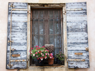 Fototapeta na wymiar Colorful daisy flowers and cactus in pots placed on the window of the old stone house. Closed window with lace curtain, rusty lattice and open wooden shutters. (Provence, France)
