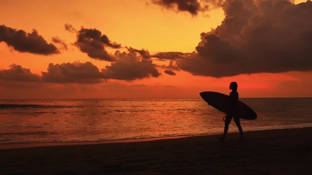 Silhouette of female surfer walking on beach and carrying surfboard at sunset with beautiful dramatic clouds on orange sky. Slow motion video of summer vacations and holidays on tropical coast od Bali