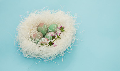 Easter eggs on white straw nest, delicate pastel color still life decoration isolated on baby blue background. Pink and green shades, white floral doodles, purple wild flower.