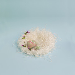 Easter eggs on white straw nest, delicate pastel color still life decoration isolated on baby blue background. Pink and green shades, white polka dots doodles, purple wild flower.