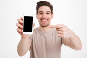Young man in casual t-shirt demonstrating mobile phone and pointing finger on copyspace screen with pleasure, isolated over white wall