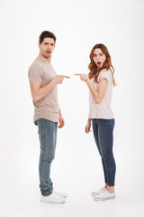 Full length photo of outraged adult guy and girl wearing beige t-shirts standing face to face and pointing fingers to each other in blaming manner, over white background