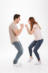 Full length photo of attractive active couple guy and girl wearing beige t-shirts expressing delight standing face to face and clenching fists, over white background