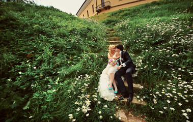 Bride and groom sitting on steps in chamomile flowers old yellow castle background at wedding Day summer nature. Bridal couple, Happy Newlywed woman and man embracing in green park. Honeymoon