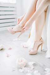 Obraz na płótnie Canvas Young woman puts on pink high heel shoes in bright room with rose flower buds. Wedding bridal fashion concept.