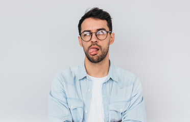 Portrait of stylish bearded male wears round trendy glasses and blue shirt, has disgusted expression with tongue. Human facial expressions, emotions, people and feelings concept