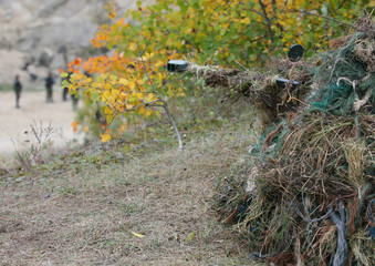 Camouflage Hunter or soldier aiming hiding in bushes in camouflage autumn  background. Sniper with rifle.