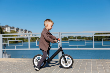 Happy four years old boy riding bicycle without pedals on the background of river and buildings. Sport concept. First balance bike for little children. Active and fun childhood outdoors