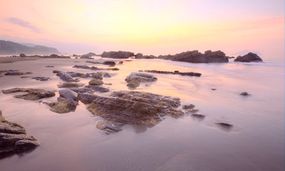 Obraz premium Sunrise scenery of a beautiful rocky beach on northern coast of Taiwan with an island on distant horizon under dramatic dawning sky & golden sunlight reflecting on the seawater (Long Exposure Effect)