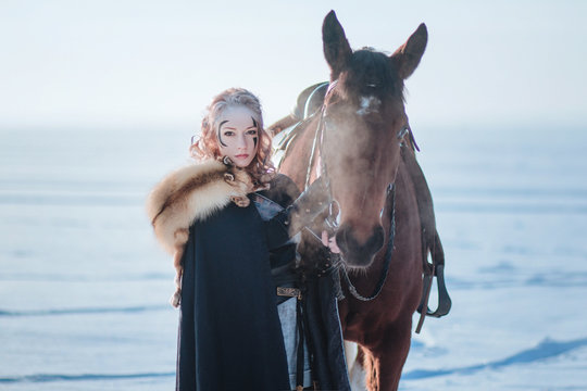 Beautiful blonde girl near the horse. Steam from the nose. Model in a warm cloak, cape. Fantasy image. Illustrations for the cover. Winter photo shoot. A lot of snow.
