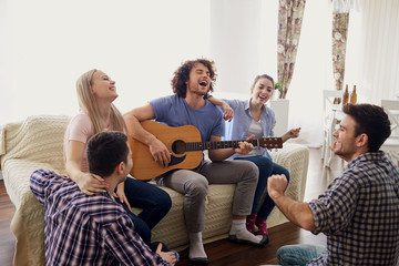 A group of friends with a guitar sing fun songs at a party indoor.