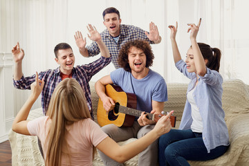A group of friends with a guitar sing fun songs at a party indoor.