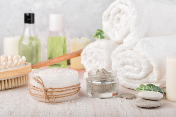 Obraz na płótnie Canvas Spa composition on white wooden background. Sea salt, white rolled towels, candles, green herbs, natural clay mask for face and body. Lotion and shampoo