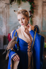 Beautiful girl in a medieval dress in blue. A model with white hair, a hairstyle with feathers. Bird of the falcon on the hand. Fantasy Photo