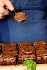 Brownie square pieces and a confectioner wearing navy blue apron is covering cakes with cacao powder