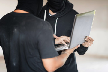 Two black masked hackers are using laptop in them hand. They are trying to  hack data. Selective focus at far hacker' hand.