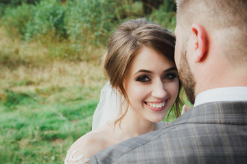 Beautiful wedding couple in the forest. The bride in tulle veil is smiling to her bearded groom in bow tie. Wedding buttonhole and checkered suit. Stylish and rustic summer love story outdoors.