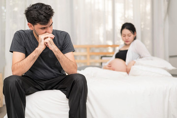 Close Up Of Serious Man Sitting On Bed With Blur Of Unhappy Pregnant Woman With Big Belly.Needless Pregnancy And Motherhood Moment People And Expectation. Family planning Concept.In Selective Focus.