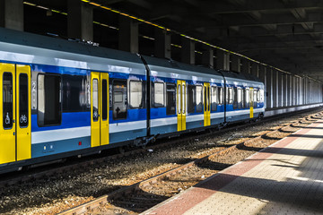 Fototapeta premium Railway station with train and platform. Covered old railway station with a modern blue and yellow train at sunny day.