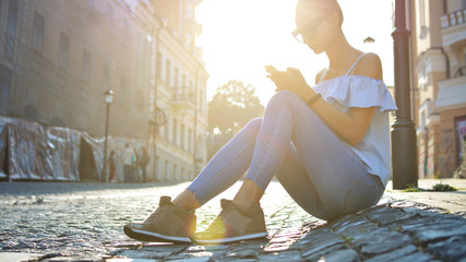 Beautiful young girl sitting on a paving stone of a European street using her smartphone. A woman communicates via the Internet. Life style. Trend. Hipster. Brunette. Sunny highlight