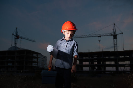 sad little caucasian boy builder in orange helmet with tool box and paper plan on construction site background