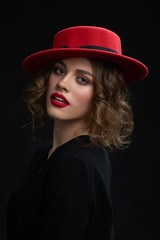 Portrait of beatiful young girl ,wering red lipstick,day make up and fashionable red hat. Model having brown curly hair and looking at camera. frontview on dark studio background.