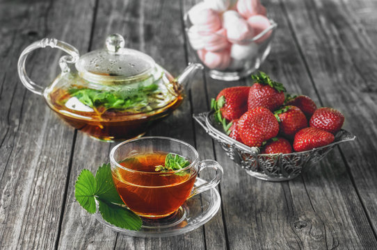 Delicious green mint strawberry tea in a transparent glass teapot and Cup in the summer, on a rustic wooden table with strawberries and marshmallows.