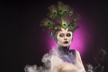 Beautiful big breast girl wearing peacock feathers in her hair and artistic violet shiny body art on her neck, vanguard makeup in theatrical smoke.