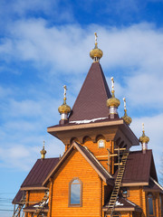 Golden domes of a wooden Church against a blue sky with clouds.
