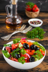 Vegetable salad with fresh tomatoes, cucumbers, black olives, red onion and parsley in a white bowl on the wooden brown table.