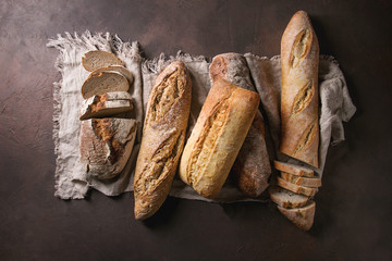 Variety of loafs fresh baked artisan rye, white and whole grain bread on linen cloth over dark brown texture background. Top view, copy space.