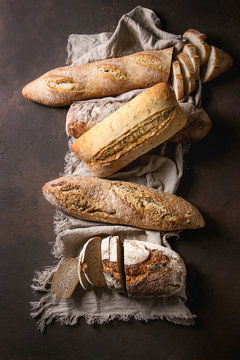 Variety of loafs fresh baked artisan rye, white and whole grain bread on linen cloth over dark brown texture background. Top view, copy space.