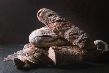 Variety of loafs fresh baked artisan rye and whole grain bread on linen cloth over dark brown texture background. Copy space.