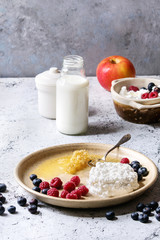 Ceramic plate of homemade cottage cheese served with blueberries, raspberries, bottle of milk and honeycombs over white marble texture table as background.