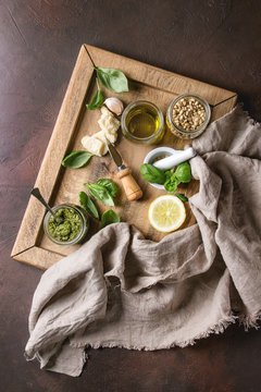 Traditional Basil pesto sauce in glass jar with ingredients above fresh basil, olive oil, parmesan cheese, garlic, pine nuts, lemon on wooden tray over dark texture background. Top view, space