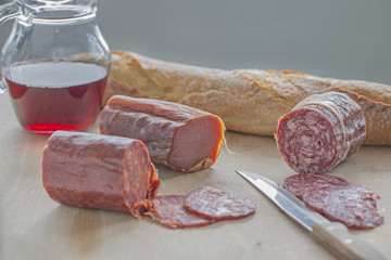 Three salami with some slices, a knife, some bread and some red wine.