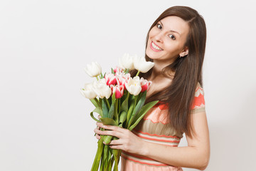 Beautiful young smiling brunette woman in light patterned dress is holding bouquet of white and pink tulips in hands, smiling and rejoices isolated on white background. Concept of holiday, good mood.